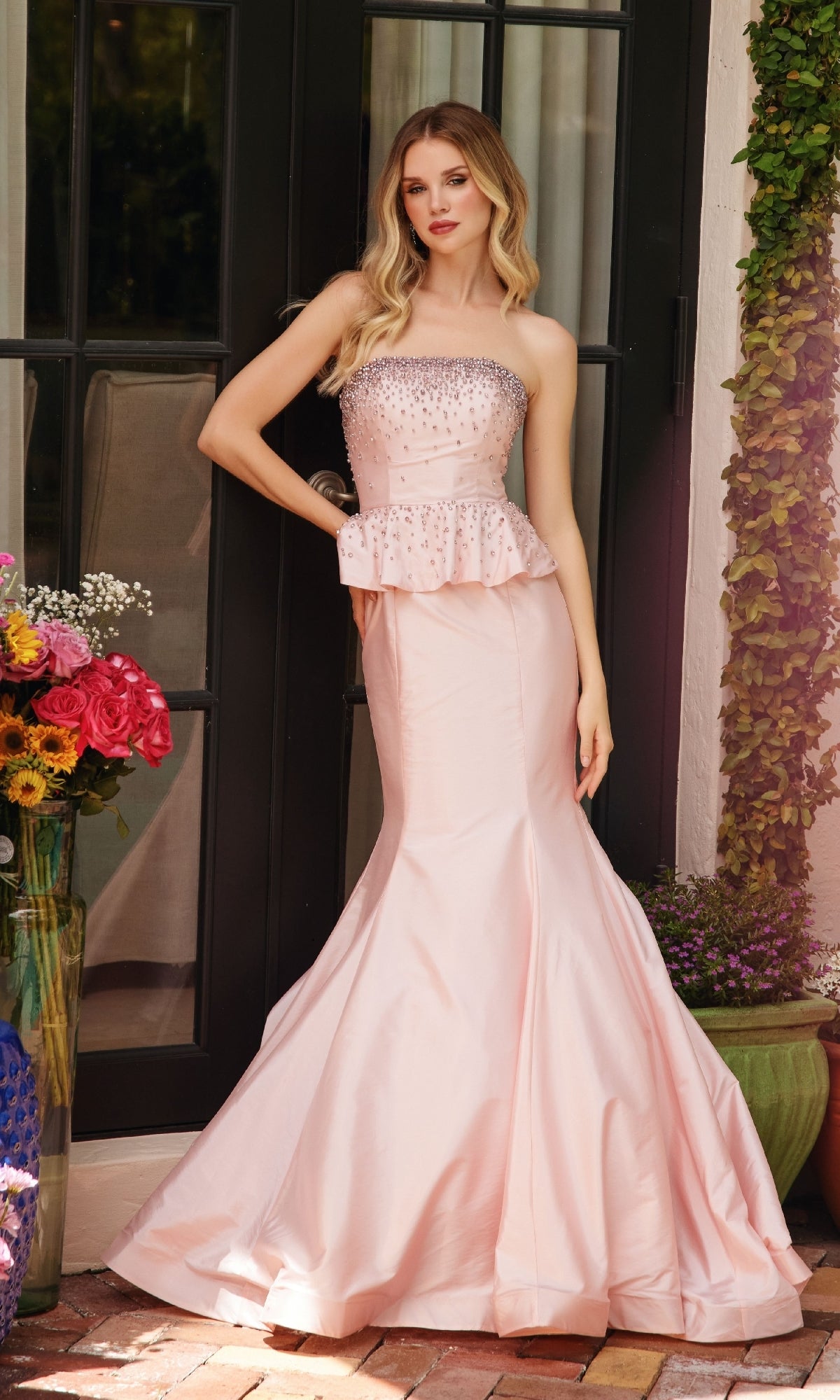 A woman stands before glass doors wearing the Ava Presley 40092 strapless peplum prom dress in light pink with beaded details. Nearby, flowers are arranged in vases.