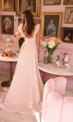 A woman in a long, light pink Ava Presley 40095 A-line prom dress stands with her back to the camera in a vintage-style room decorated with floral arrangements, tea sets, and portraits on pink walls.