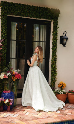 Wearing the Ava Presley 40095 Long A-Line Prom Dress, a woman with ruffle shoulder straps stands before black French doors, surrounded by potted flowers and brick flooring in an outdoor setting.