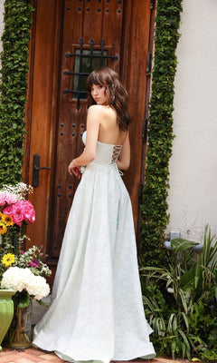A woman in the Ava Presley 40097 Rosette A-Line Prom Dress poses on a brick doorstep surrounded by flowers and greenery, facing away from the camera. The dress is strapless and light blue.