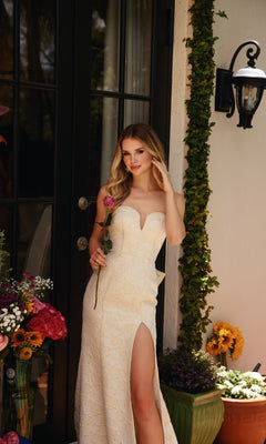 Wearing the Back-Ruffle Textured Prom Dress: Ava Presley 40098, a woman stands outdoors with a single rose, framed by lush flower arrangements and greenery.