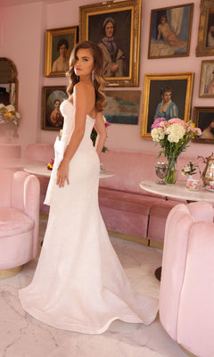 A woman wearing the Ava Presley 40099 long lace prom dress with a bow stands in a pink room with antique portraits, pink velvet chairs, and a table topped with a flower bouquet.