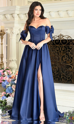 A woman wears a floor-length navy blue Colors Dress 4009 A-line prom dress with an off-the-shoulder sweetheart neckline, ribbon sleeve details, and a high side slit.