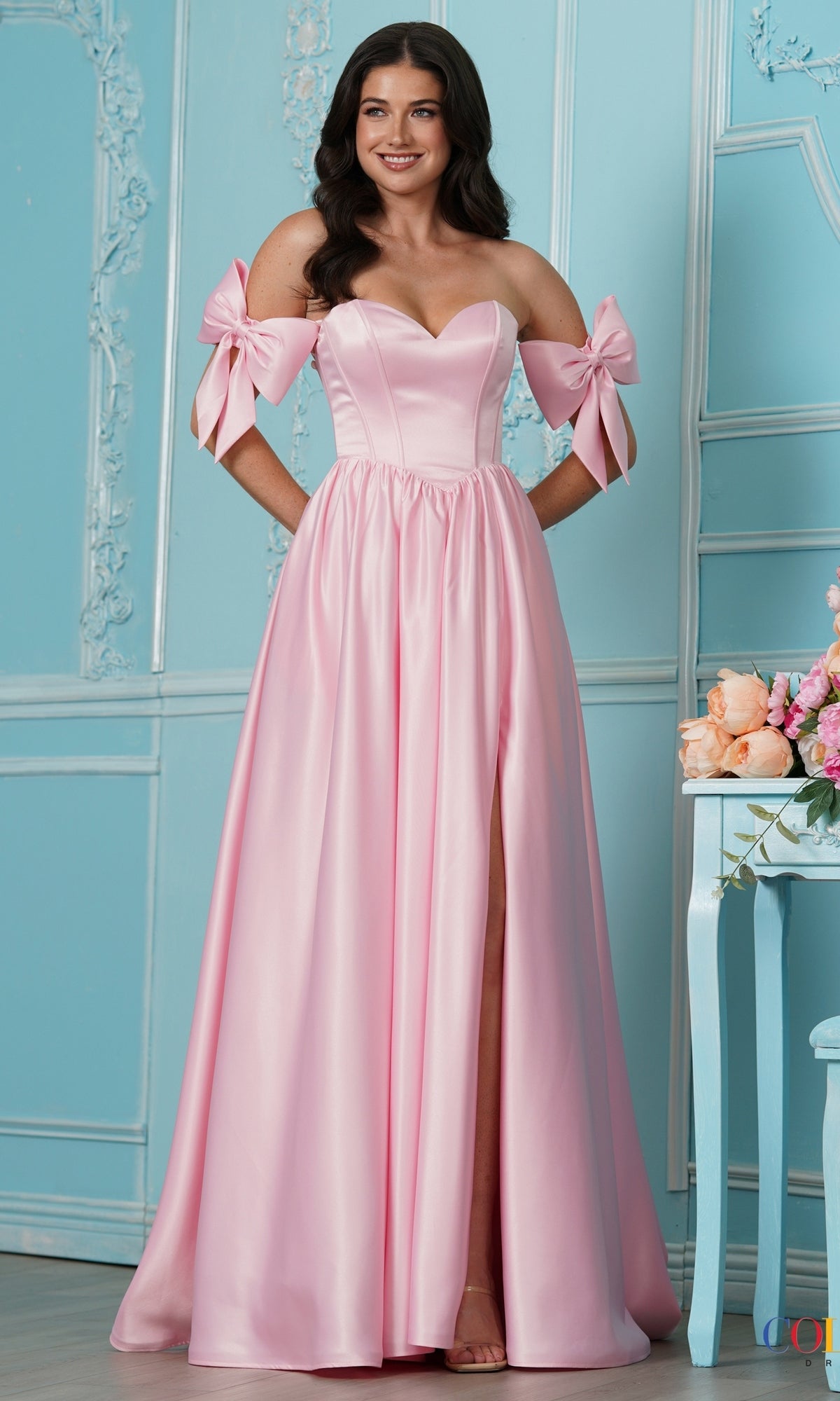 A woman wearing a pink, floor-length off-the-shoulder bow Long Prom Dress: Colors Dress 4009 with a high leg slit.