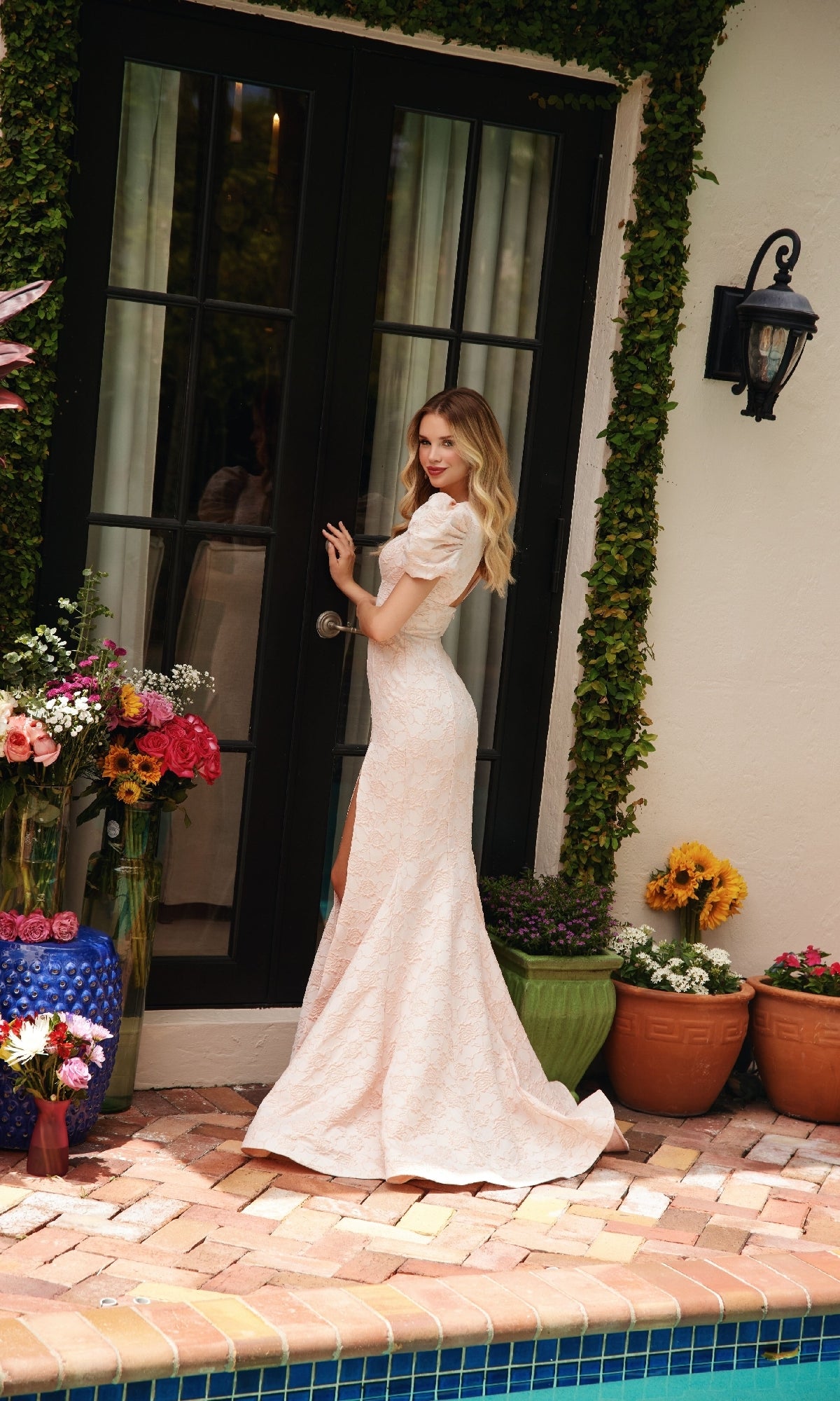 A woman wearing the Ava Presley 40100 V-Neck Puff-Sleeve Prom Dress stands by glass doors, surrounded by potted plants and vibrant flower arrangements near a pool.