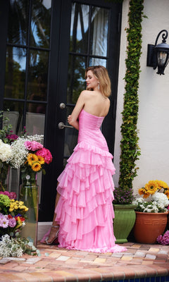 A woman wearing the Strapless Long Ruffle Prom Gown Ava Presley 40118 stands by a glass door on a tiled patio, surrounded by potted flowers and plants.
