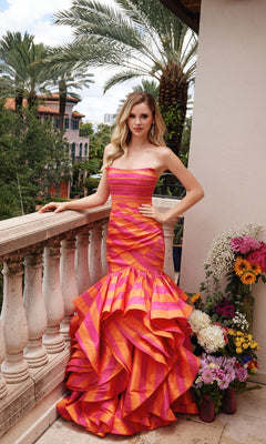 A woman wearing the Ava Presley 40119 long formal dress, a strapless mermaid gown with ruffled orange and pink layers, stands on a balcony beside flowers, with palm trees and city buildings in the background.