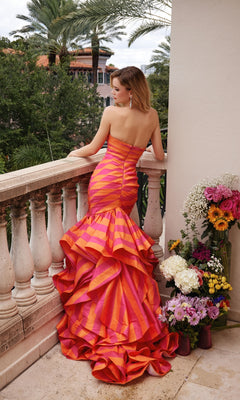 A woman wearing the Ava Presley 40119 long formal dress, featuring a strapless mermaid silhouette with pink and orange ruffles, stands on a balcony by vibrant flowers with palm trees in the background.