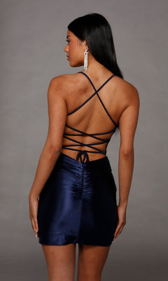 A woman stands with her back to the camera in a navy blue Alyce 40123 Lace-Up Short Satin Sheath Hoco Dress, featuring crisscross straps and ruched detailing, set against a plain gray background.