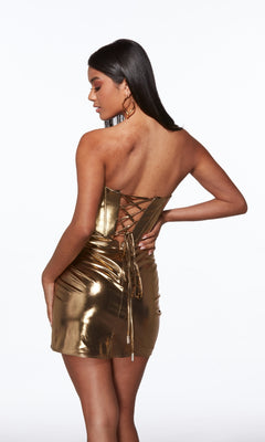 A woman stands with one hand on her hip, facing away from the camera in the Alyce 40124 short homecoming dress, featuring a metallic strapless design with a corset bodice and lace-up open back.