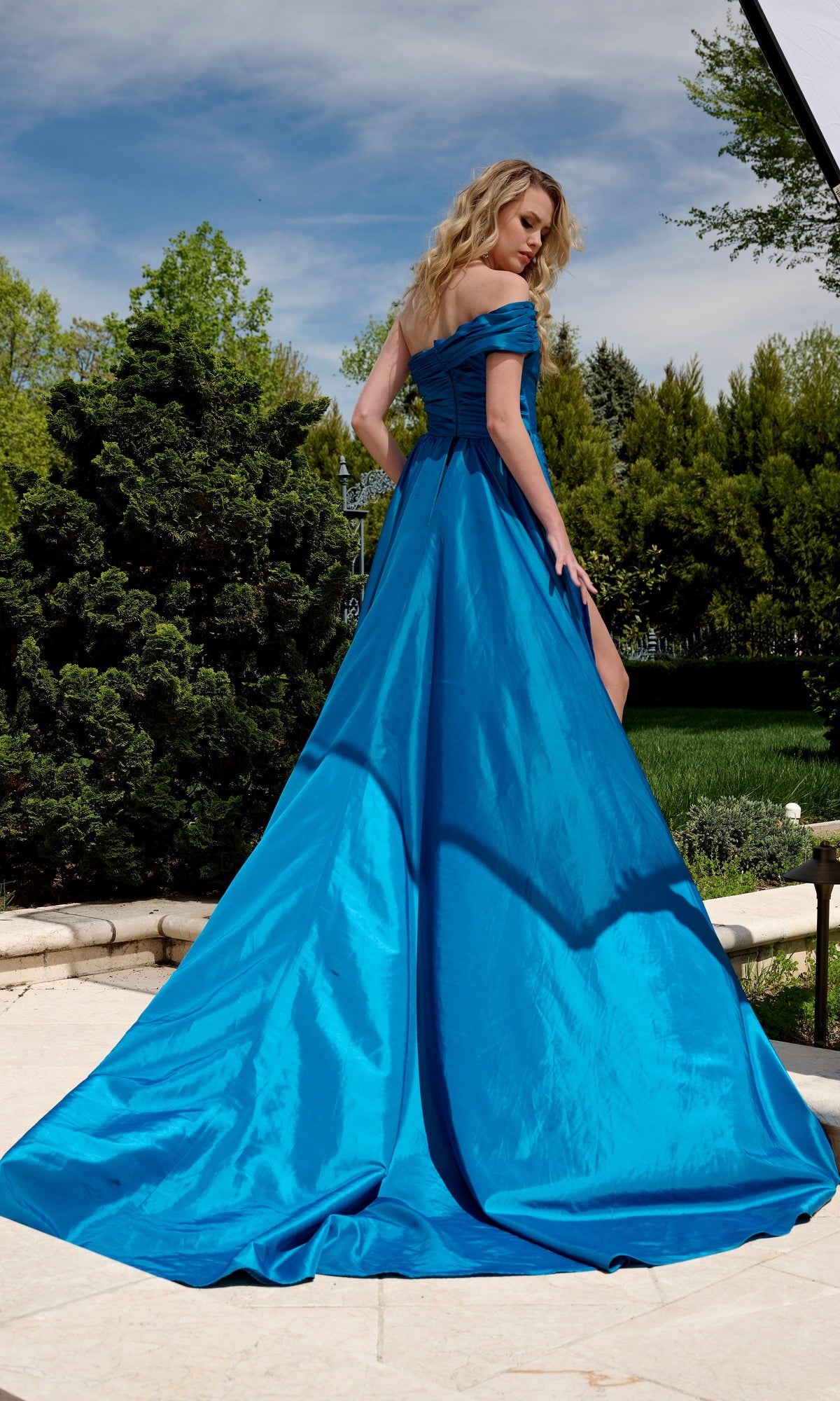 A woman stands outdoors on a stone patio in the Jovani 40128 long prom dress, featuring an off-the-shoulder design and thigh-high slit. The bright teal-blue gown contrasts beautifully with the lush green trees behind her.