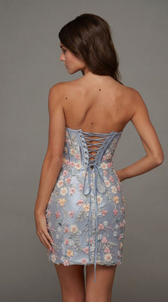 A woman facing away is wearing the Alyce 40139 short homecoming dress, which features a strapless design with a lace-up back and corset bodice. The light blue fabric is adorned with pastel floral embroidery and appliques. Neutral background.