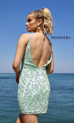 A woman with blonde hair in a ponytail wears the Low Open-Back Mini Beaded Party Dress 4013—a light green short dress—standing by the ocean under a clear blue sky with “PRIMAVERA” displayed in the background.