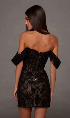 A woman facing away models the Short Homecoming Dress: Alyce 40142—a black, off-the-shoulder sequin dress featuring a fitted silhouette, visible zipper, and sheer lace detailing—capturing its timeless elegance against a plain gray background.