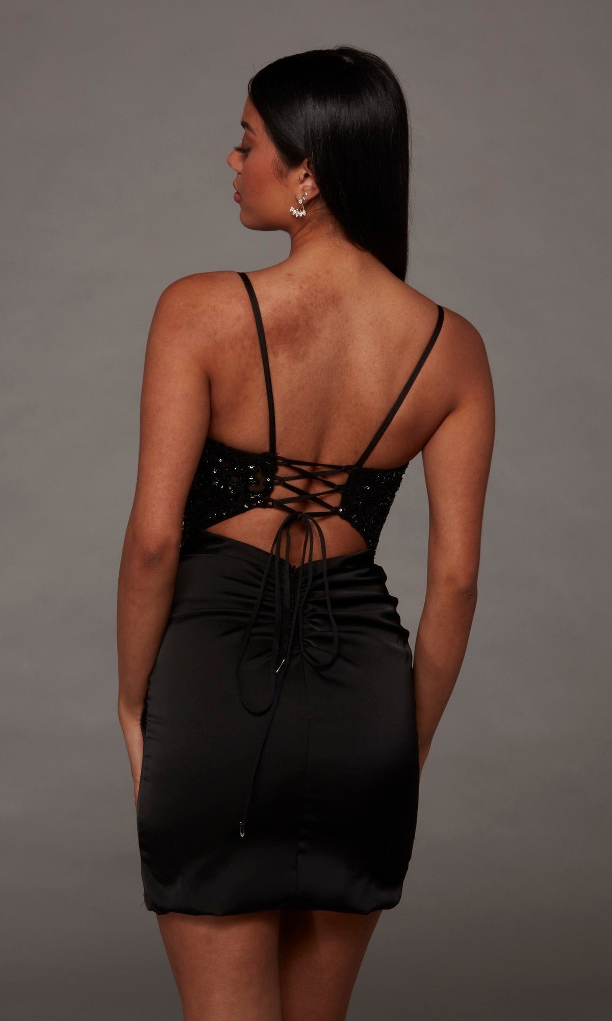 A woman stands facing away, highlighting the lace-up detailing and strappy, backless design of the Alyce 40143 Short Homecoming Dress. Her dark straight hair contrasts with the gray background, while the sequin-embellished bodice lends subtle shimmer to this elegant silhouette.