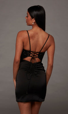 A woman stands facing away, highlighting the lace-up detailing and strappy, backless design of the Alyce 40143 Short Homecoming Dress. Her dark straight hair contrasts with the gray background, while the sequin-embellished bodice lends subtle shimmer to this elegant silhouette.
