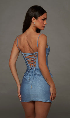 A woman is seen from behind wearing the Alyce 40146 short homecoming dress, featuring a sparkly light blue fabric, lace-up back, and corset bodice, standing against a plain gray background.