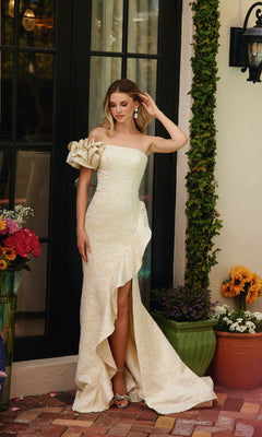 A woman in the Ava Presley 40146 Ruffle Flower Prom Dress, featuring an off-shoulder cream design with a ruffled sleeve and high slit, stands outside by a glass door, surrounded by potted flowers.