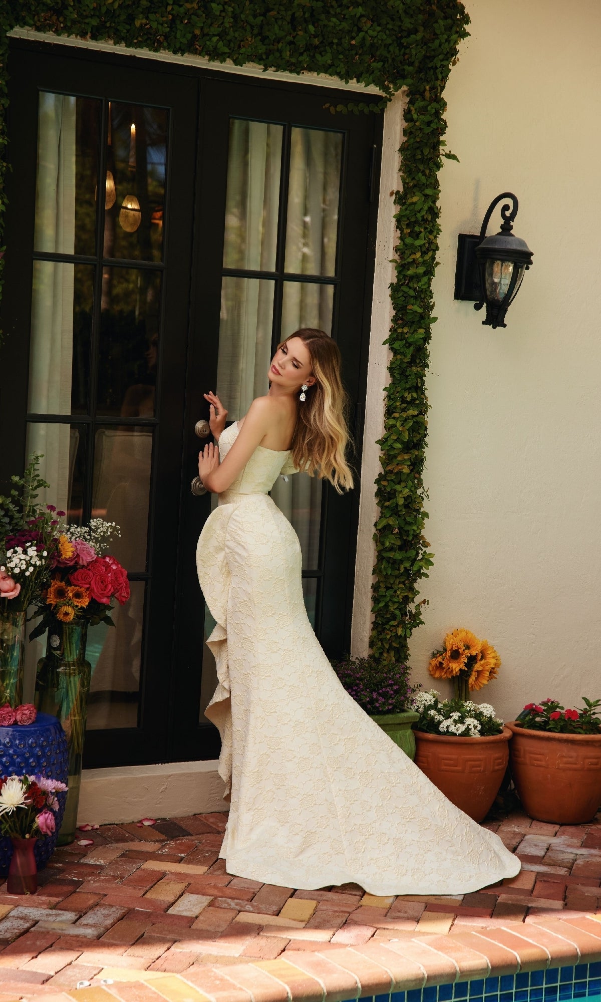 Woman in a strapless, off-white Ruffle Flower Prom Dress: Ava Presley 40146 stands by a glass door surrounded by potted flowers and greenery, looking over her shoulder.