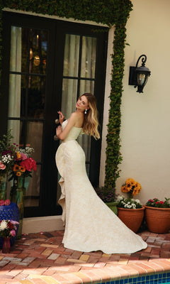 Woman in a strapless, off-white Ruffle Flower Prom Dress: Ava Presley 40146 stands by a glass door surrounded by potted flowers and greenery, looking over her shoulder.