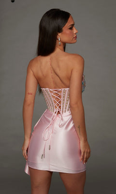 A woman stands facing away, showcasing her long dark hair, a visible back scar, the number “444” tattooed on her right arm, and small tattoos on her left arm. She wears the Short Homecoming Dress: Alyce 40147—a shiny light pink mini dress featuring a strapless lace-up corset bodice and adorned with delicate 3D floral appliques for an extra touch of elegance.