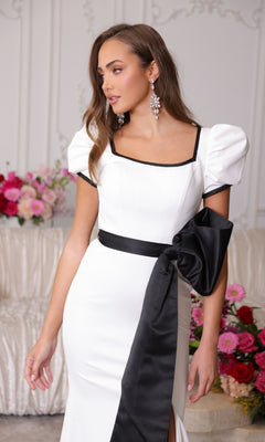Woman wearing the Ava Presley 40152 long prom dress, featuring a form-fitting silhouette, black trim, puff sleeves, and a large black bow, posing indoors with floral arrangements in the background.