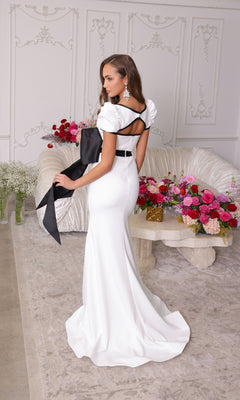 Wearing the Ava Presley 40152 long prom dress, a woman stands in an elegant room with floral arrangements and ornate walls. The form-fitting gown features puff sleeves and a leg slit for a stunning look.