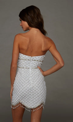 A woman faces away, wearing the Alyce 40153 short homecoming dress—a strapless white mini dress featuring delicate pearl-embellished beads, intricate beading, and a scalloped hem. Gray background.
