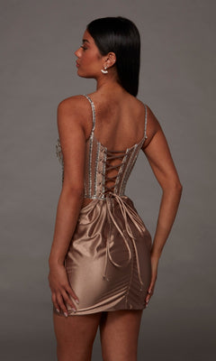 A woman stands with her back to the camera, modeling the Alyce 40160 Short Homecoming Dress—a shiny tan mini dress featuring lace-up detailing and jeweled straps—set against a plain gray background. This chic dress is adorned with elegant accents that make it a standout choice for homecoming.