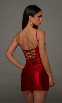 A woman stands with her back to the camera against a plain gray background, showcasing the Alyce 40162 short homecoming dress—a vibrant red satin mini dress featuring a fitted silhouette and lace-up back that exudes chic elegance for any special occasion.