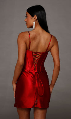 A woman facing away showcases the Alyce 40163 short homecoming dress—a shiny red satin corset dress with lace-up back and spaghetti straps—set against a plain gray background, making it perfect for an unforgettable mini homecoming look.