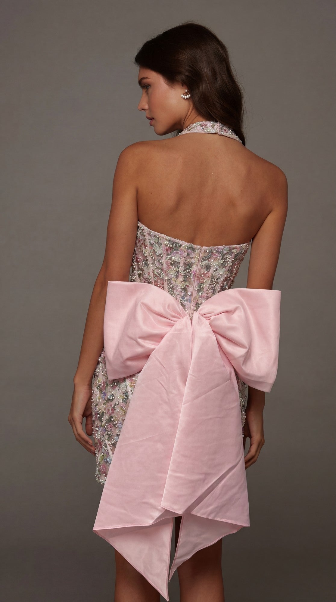 A woman wearing the Short Homecoming Dress: Alyce 40165, which features a strapless design with 3D sequin floral embroidery and a large pink bow at the back, stands against a plain gray background, turned slightly to the side.