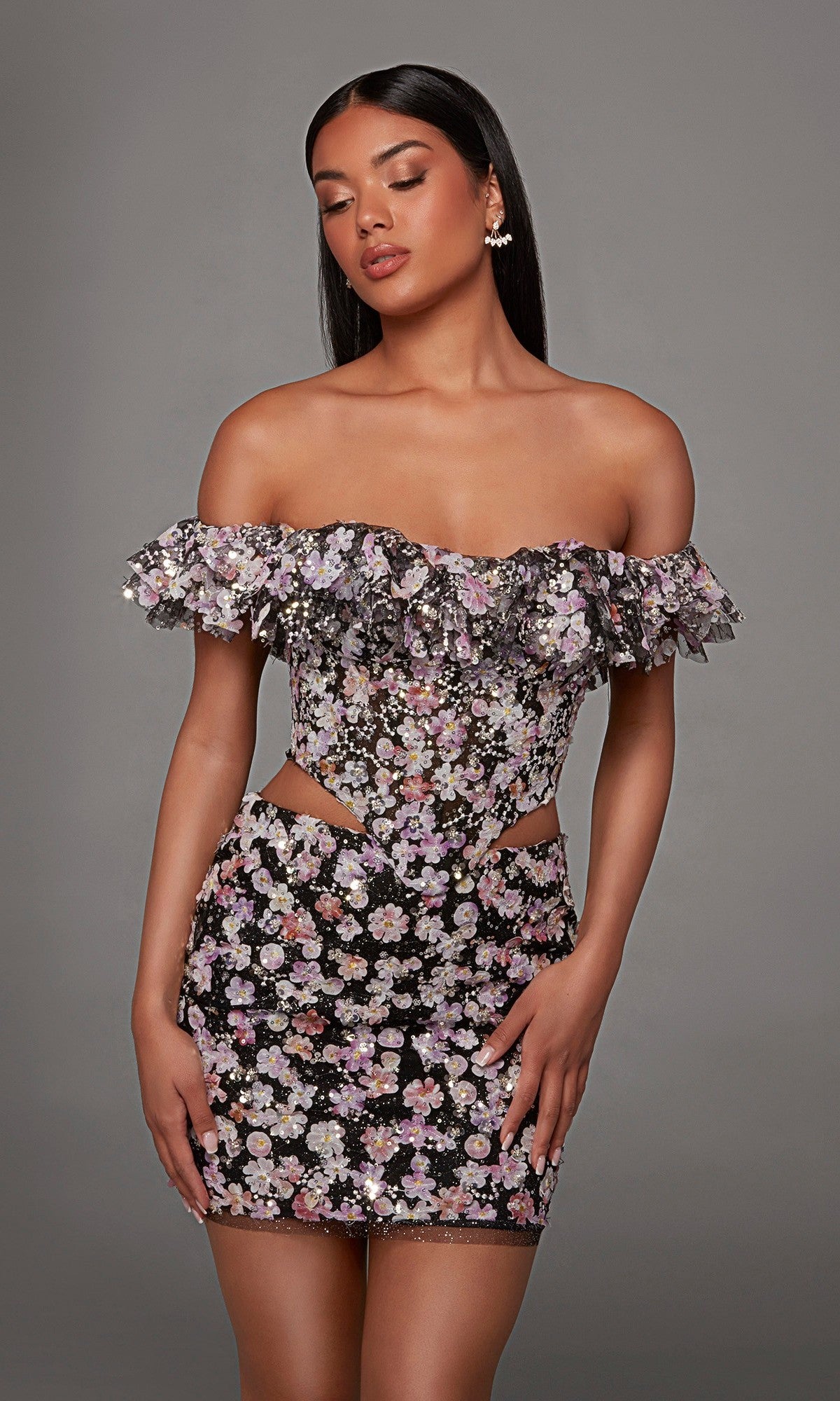 A woman poses against a plain gray background wearing the Alyce 40167 short homecoming dress in Black/Multi, featuring an off-the-shoulder neckline with ruffles, a fitted silhouette, floral print with 3D embroidered appliques on the cutout sides.