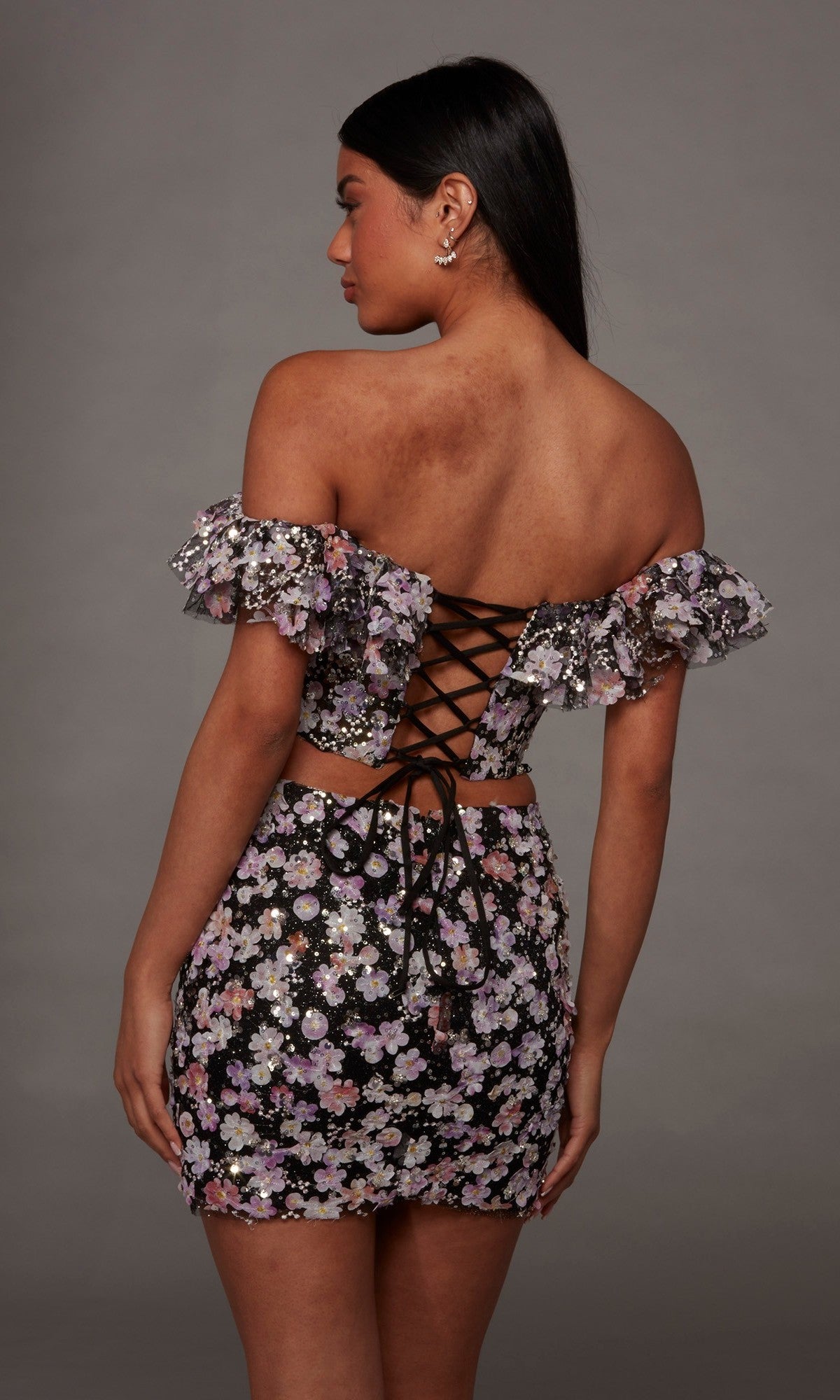 A woman faces away, wearing the Alyce 40167 Short Homecoming Dress—an off-shoulder crop top and matching skirt adorned with delicate 3D floral embroidered appliqués and a lace-up back—standing against a plain gray background.