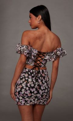 A woman faces away, wearing the Alyce 40167 Short Homecoming Dress—an off-shoulder crop top and matching skirt adorned with delicate 3D floral embroidered appliqués and a lace-up back—standing against a plain gray background.