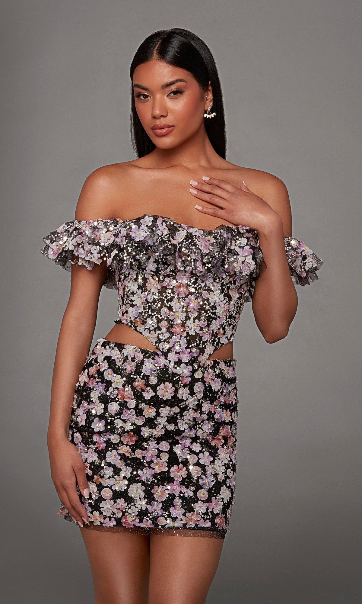 A woman poses in the Alyce 40167 short homecoming dress, showcasing an off-shoulder black design with delicate 3D floral embroidered appliques, frilled sleeves, and side cutouts, set against a plain gray background.
