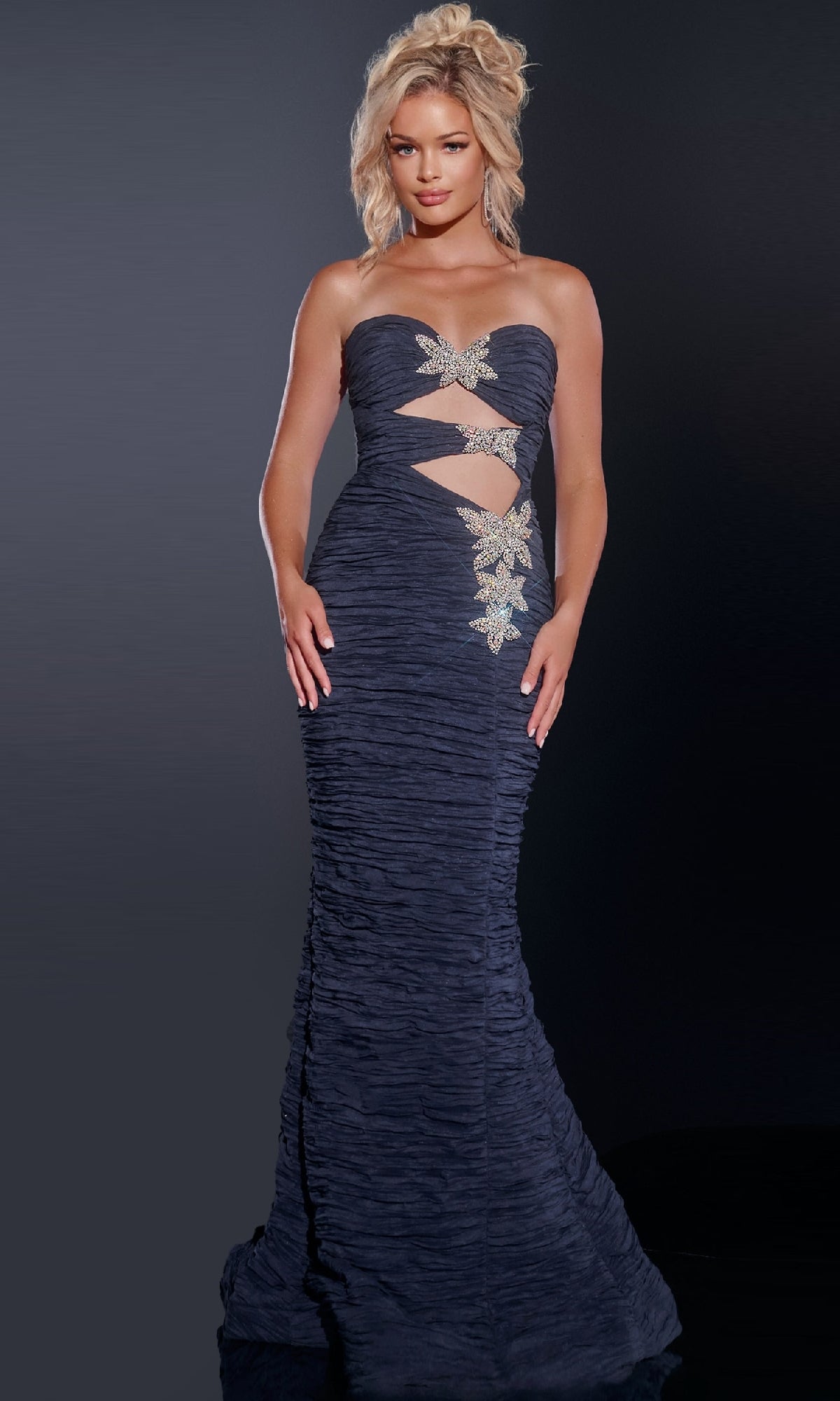 A woman wears the Jovani 40174 long prom dress, featuring a strapless design, ruched detailing, front cutouts, and floral embellishments, standing against a dark background.