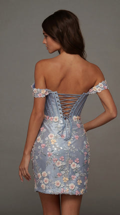 A woman stands with her back to the camera, wearing the Alyce 40175 short homecoming dress—a light blue stretch-satin style featuring 3D floral embroidery and a lace-up corset back.
