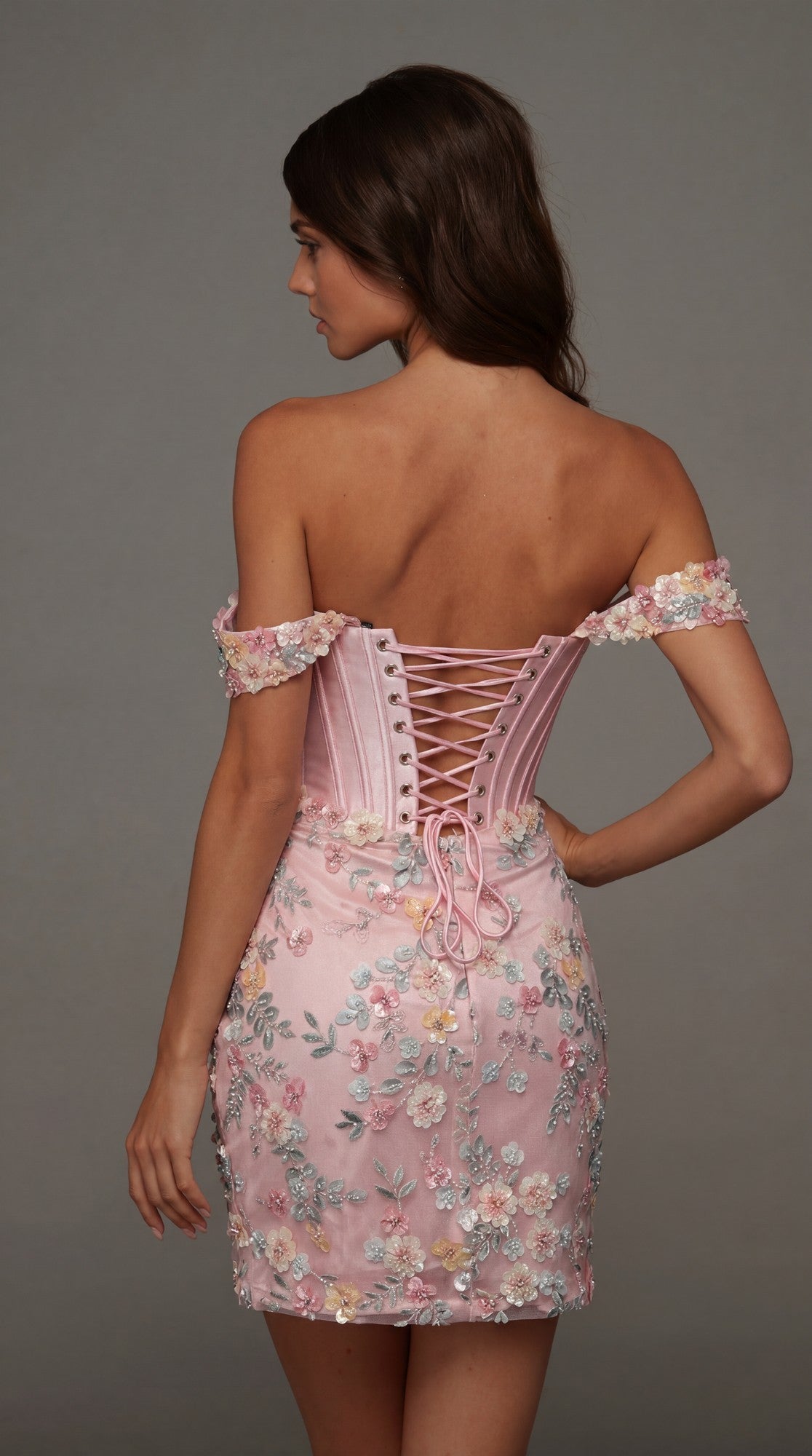 A woman faces away, modeling the Alyce 40175 short homecoming dress—a pink off-shoulder style with lace-up back, 3D floral embroidery, and short sleeves—standing against a plain gray background.