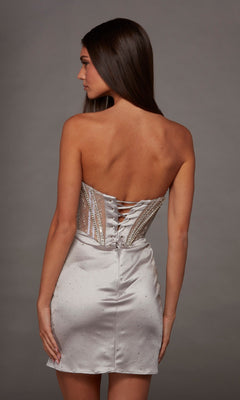 A woman in the Alyce 40193 short homecoming dress, a strapless silver mini with rhinestone accents and a lace-up back, stands against a gray background facing away from the camera.
