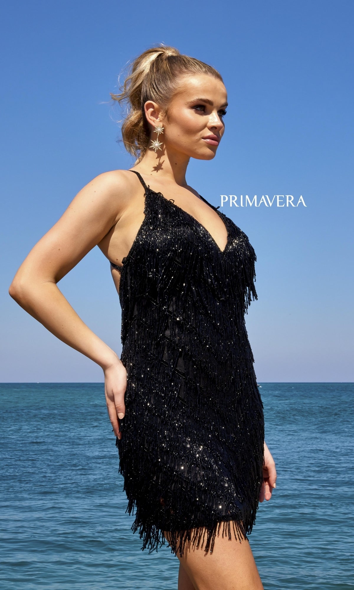A woman in the Beaded Fringe Open-Back Short Party Dress 4019 stands by the ocean under a clear sky, with 
