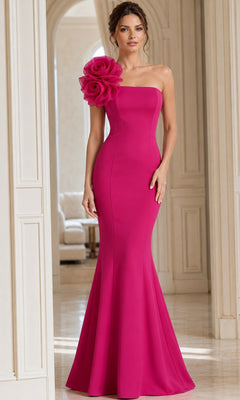 A woman wearing the Long Formal Dress: Jovani 40201, a strapless, fitted fuchsia designer gown featuring a large floral detail on the left shoulder, stands in an elegant, light-filled room.