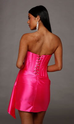A woman wearing the Alyce 40207 short homecoming dress in bright pink stretch-satin, showcasing its strapless design and corset bodice with lace-up back, stands against a plain gray background, facing away from the camera.