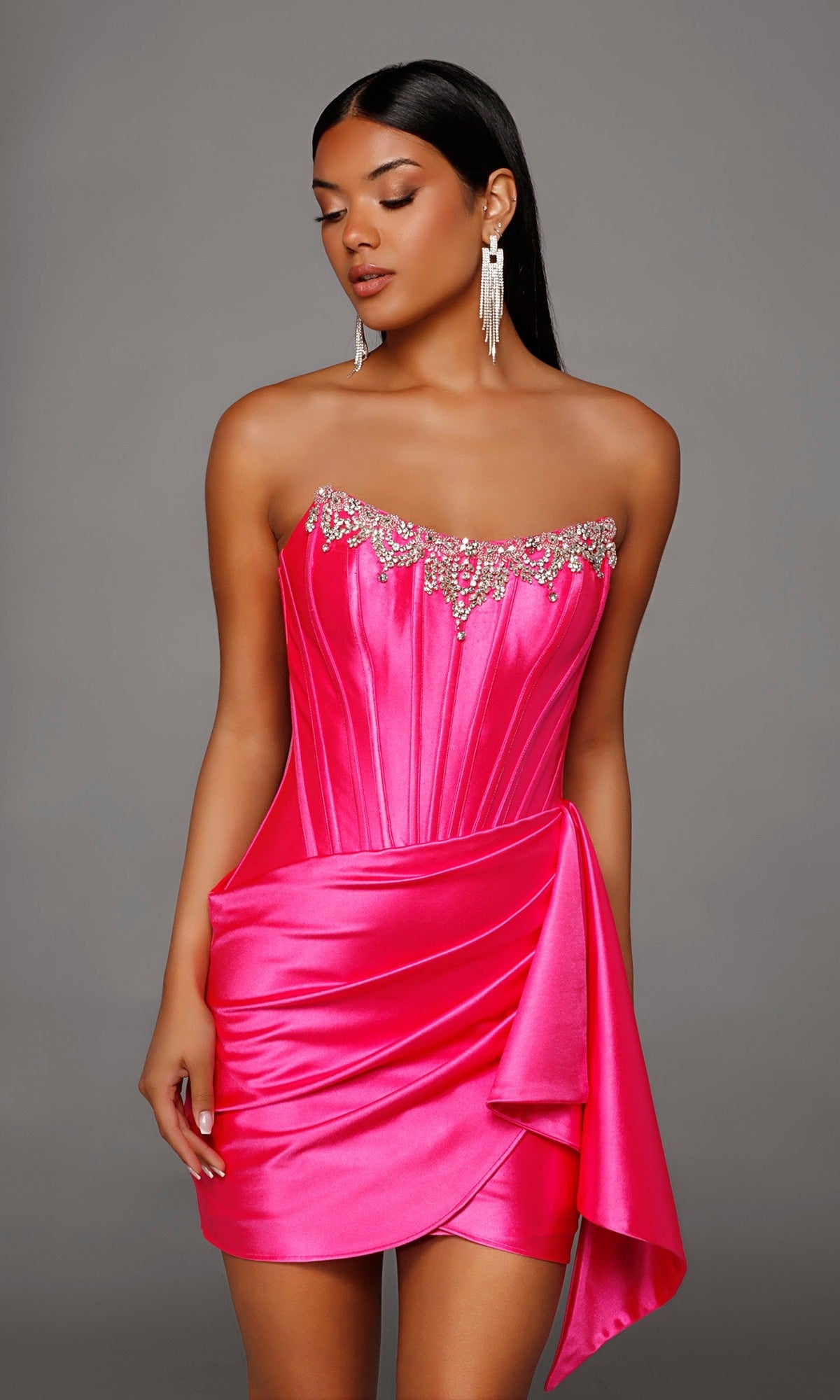 A woman stands against a plain gray background wearing the Alyce 40207, a short homecoming dress crafted from bright pink stretch-satin with a strapless corset bodice embellished with rhinestone detailing at the top and draped fabric accenting the skirt.