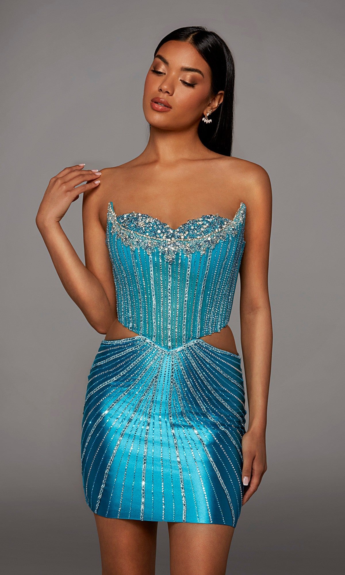 A woman wearing the Alyce 40211 Short Homecoming Dress in Lagoon Blue—a strapless mini dress featuring a corset bodice, rhinestone embellishments, and side cutouts—poses against a plain gray background.