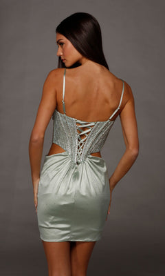 Woman wearing the Alyce 40218 short homecoming dress, a light green stretch-satin mini dress with a fitted skirt, embellished corset bodice, lace-up back, and spaghetti straps, standing against a gray background—an elegant choice for your next homecoming event.