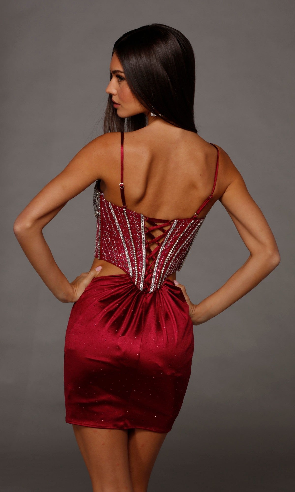 Wearing the Short Homecoming Dress: Alyce 40218—a red, sleeveless, backless dress featuring an embellished corset bodice, lace-up detail, and a fitted stretch-satin skirt—a woman stands with her hands on her hips, facing away from the camera.