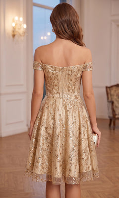 A woman with wavy brown hair stands in an elegant room, facing away from the camera, wearing the Chicas 4021 Short Homecoming Dress—an off-the-shoulder, sweetheart neckline, knee-length dress with gold embroidery.
