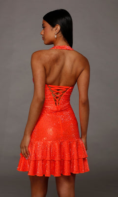 Wearing the Short Homecoming Dress: Alyce 40226, a person stands facing away to reveal the sparkly, bright orange halter style with rhinestone accents, lace-up back, ruffled skirt, and bare shoulders set against a plain gray background.