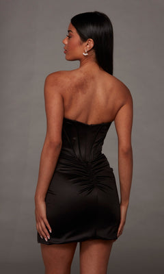 A woman with long dark hair is seen from behind, wearing the Alyce 40236 Short Homecoming Dress—a strapless black ruched mini dress with a corset bodice—against a plain gray background.